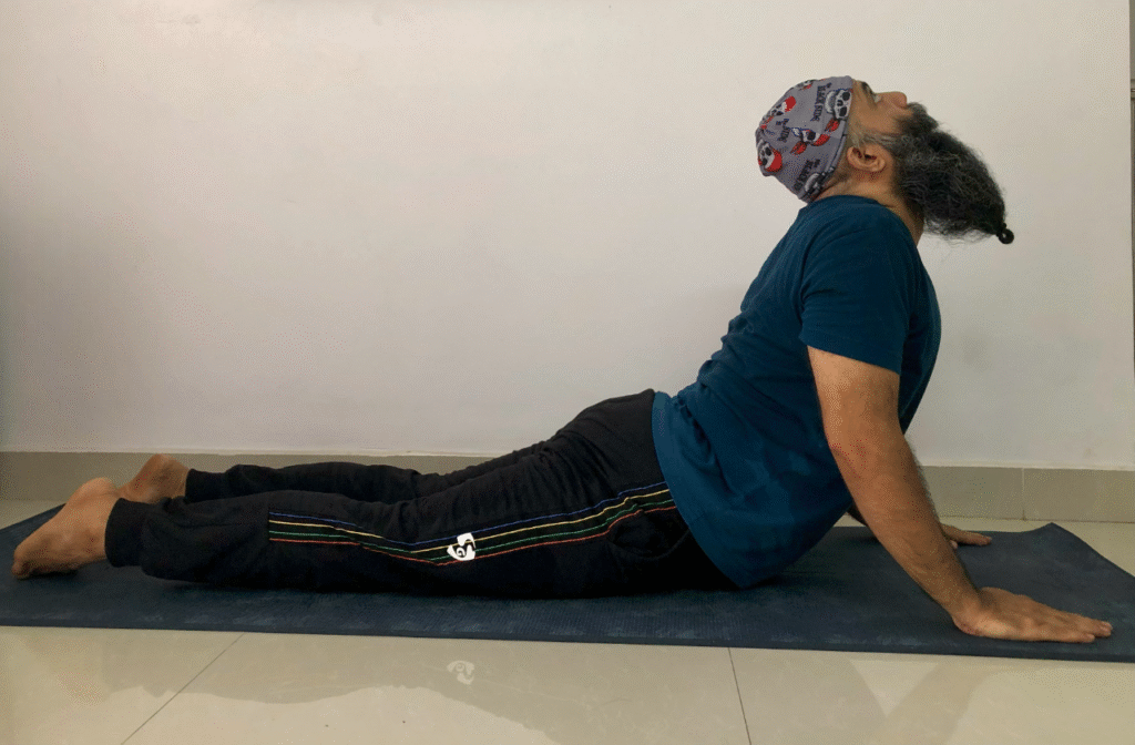 A man with a beard and cap demonstrates Bhujangasana yoga pose, lying on his stomach with his chest lifted.