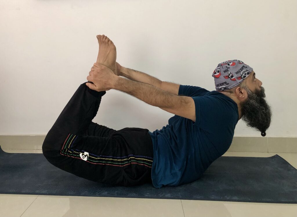 A man with a beard and hat demonstrates Dhanurasana, the yoga pose, lying on his stomach with arms raised.