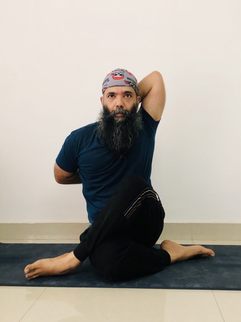 A man with a beard and cap is practicing Gomukhasana, a seated yoga pose, on a yoga mat.