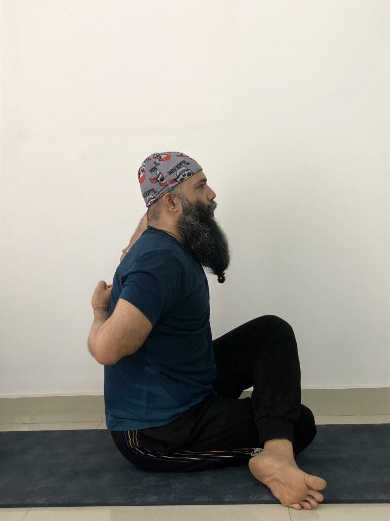 A bearded man in a cap performs Gomukhasana yoga pose, sitting cross-legged on a mat.