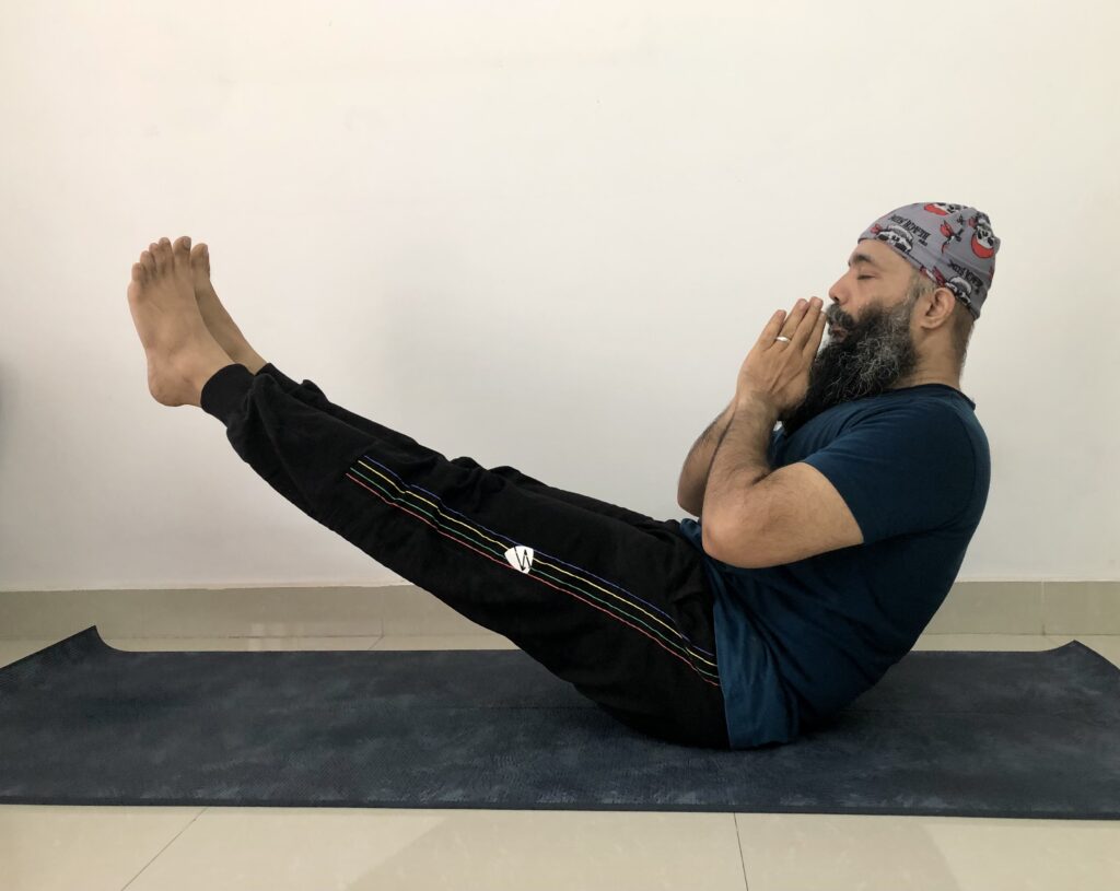  A man with a beard and cap demonstrates Naukasana yoga pose, lying on his back with legs and arms lifted.